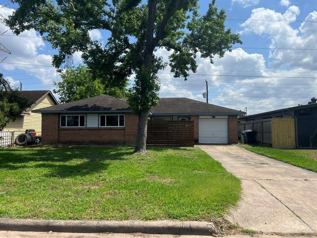 This is a single-story brick house with a one-car garage, set on a grassy lot with a large tree in the front yard. The driveway leads to the garage, and the home features a simple, welcoming facade with several windows.