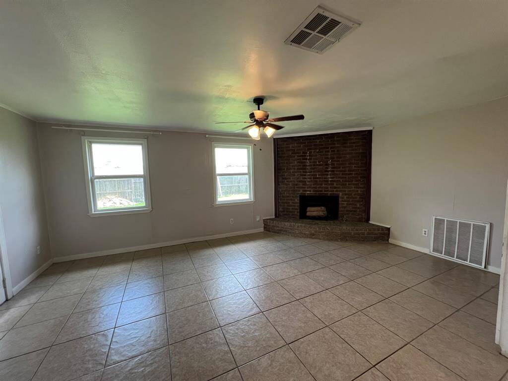Spacious living room with tiled floors, a brick fireplace, ceiling fan, and ample natural light from two windows.
