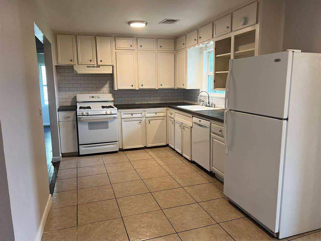 This kitchen features tiled flooring, white cabinetry, and a window above the sink. It includes a gas stove and dishwasher with a neutral backsplash and ample storage space.