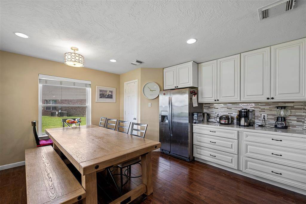A bright, inviting kitchen with white cabinetry, stainless steel appliances, large enough for a large wooden dining table with ample seating space. Some features include hardwood floors, recessed lighting, and a window overlooking a green outdoor space, creating a warm and functional dining area.