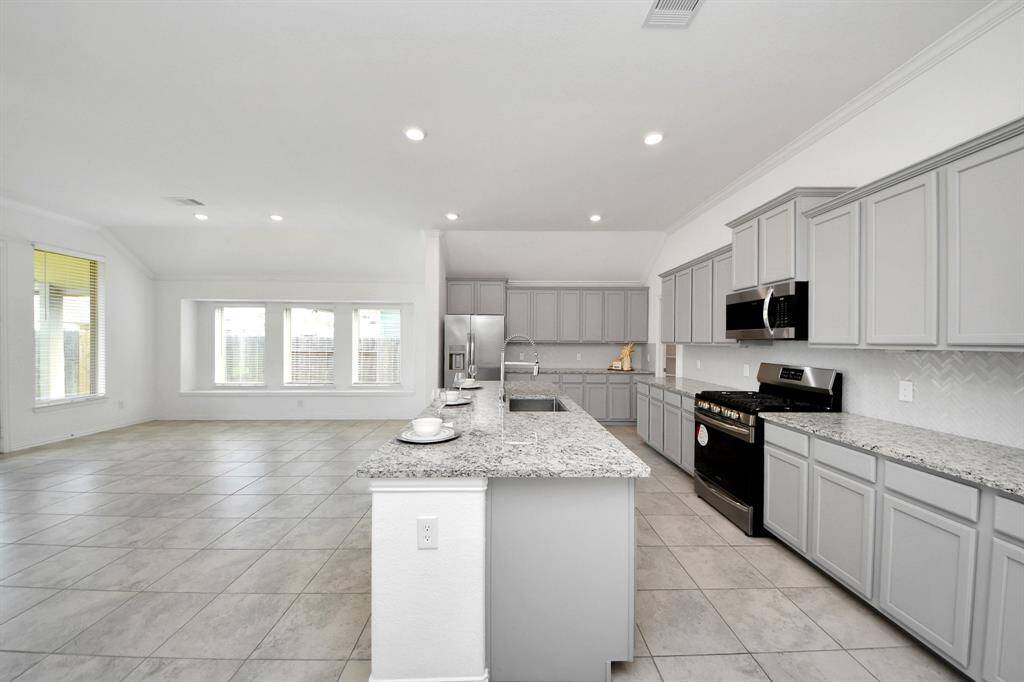 Bright and spacious kitchen with modern gray cabinets, granite countertops, and stainless steel appliances. The open layout flows into a large living area with ample natural light from multiple windows.