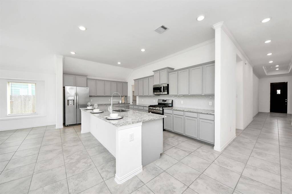 This modern kitchen features sleek light gray cabinetry, stainless steel appliances, and a large island with bar seating. The open layout and ample lighting create a spacious and inviting atmosphere, perfect for entertaining.