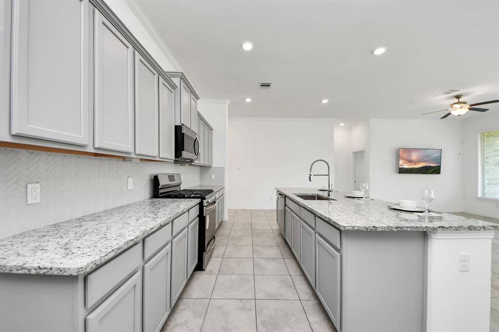 Spacious kitchen with modern gray cabinets, granite countertops, and a large island. Features stainless steel appliances, tile flooring, and an open layout leading to a bright living area with a ceiling fan.
