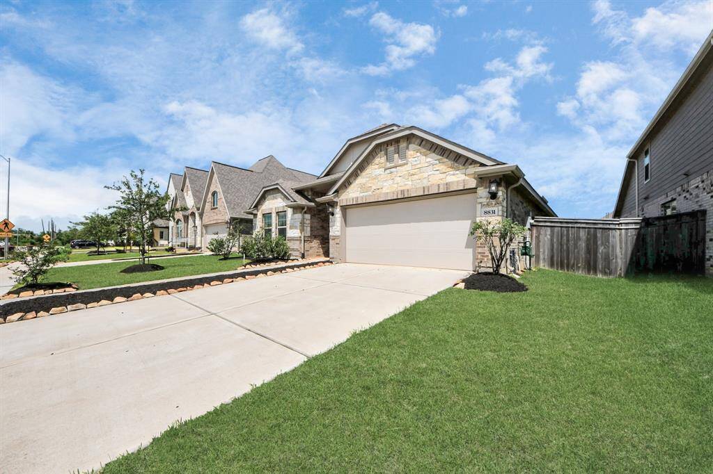 This photo showcases a modern, single-story home with a stone and brick facade, attached two-car garage, and a well-maintained front lawn. It is situated in a residential neighborhood with similar style homes and a clear blue sky overhead.