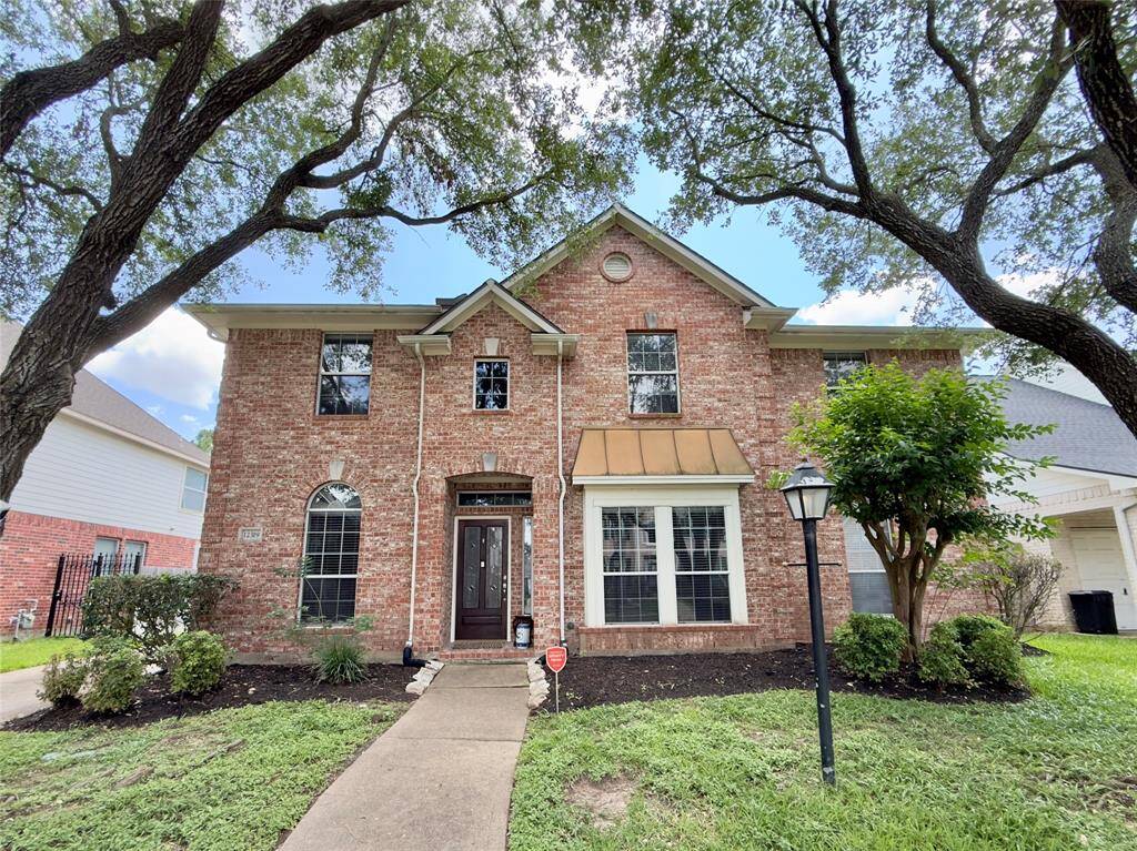 This charming two-story brick home features a welcoming front entrance with a brown door and a small porch. It is surrounded by mature trees and well-maintained landscaping, offering a cozy and inviting curb appeal.