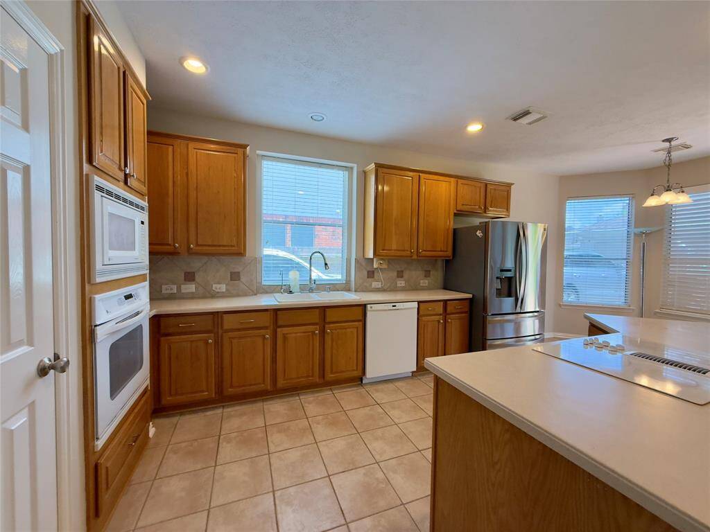This kitchen features warm wood cabinets, a stainless steel refrigerator, and a white oven and dishwasher. It has a spacious layout with ample natural light from large windows and neutral tile flooring.