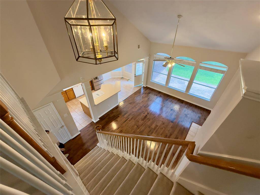 This spacious home features an open living area with high ceilings, large windows providing excellent natural light, and a view of the backyard. The room boasts beautiful hardwood floors, a stylish hanging light fixture, and an elegant staircase. The adjacent kitchen is tiled and well-lit, making it ideal for entertaining and family gatherings.