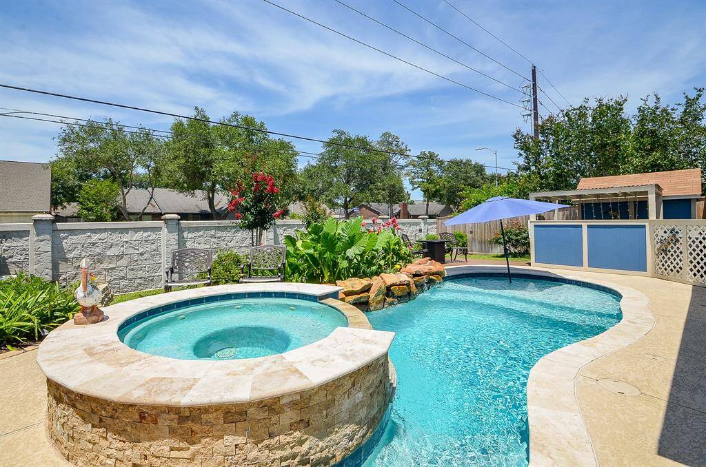 Beautiful swimming pool and spa with stone accents, surrounded by lush greenery and landscaping. A patio area with seating and an umbrella provides a perfect spot for relaxation and outdoor entertaining. The area is enclosed by a privacy fence, offering a serene and private outdoor space.