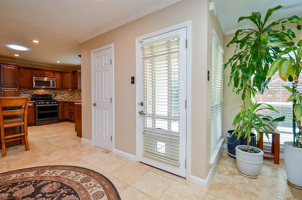 "Welcome." Entry showcases a bright, inviting kitchen area with warm wooden cabinetry, a modern stainless steel oven, and Travertine flooring. Adjacent is a cozy nook great for potted plants and a door with blinds, adding a touch of greenery and natural light.