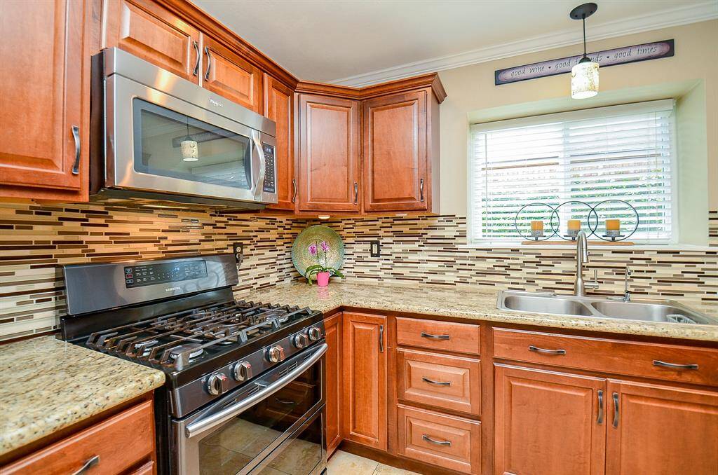 Kitchen features warm wooden cabinets, a modern stainless steel gas stove and microwave, and a double sink beneath a bright window. The stylish mosaic tile backsplash complements the granite countertops, creating a cozy and functional cooking space.