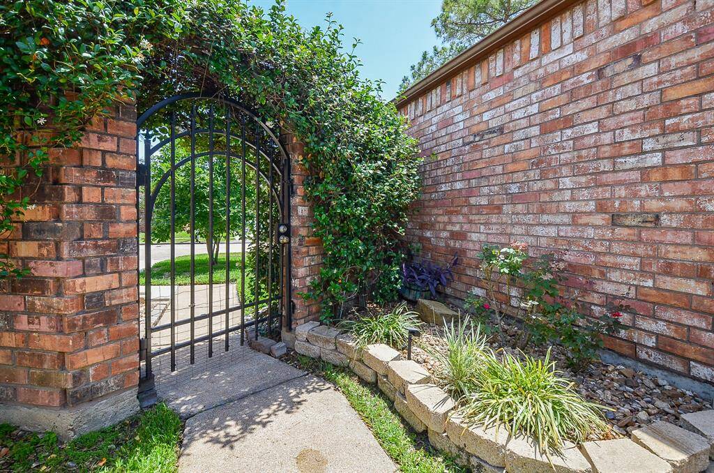 Charming brick entryway with an arched iron gate covered in greenery. A well-maintained garden bed adds curb appeal, leading to a welcoming pathway.