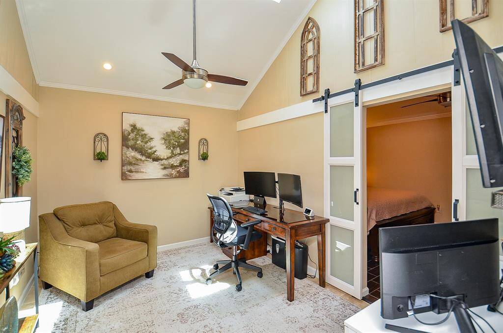 This photo showcases a cozy home office with a vaulted ceiling, neutral walls, and a ceiling fan. It features a dual sliding barn door leading to the primary suite.  Natural light enhances the space.
