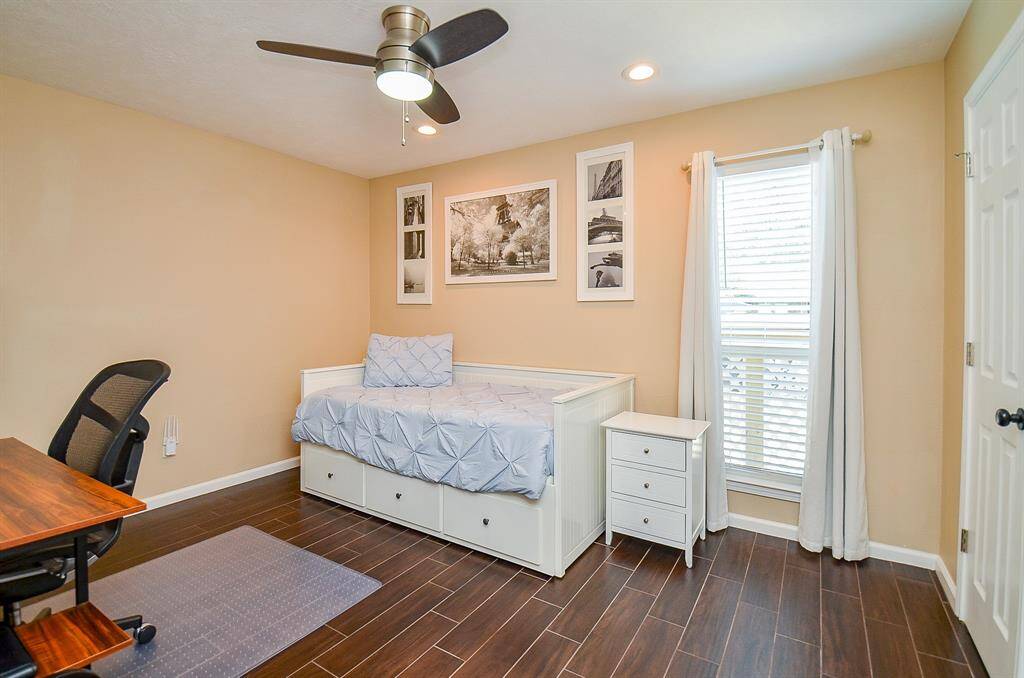 Versatile bedroom is set up for an office with room for bed.  Nice lighting with white curtains for natural light. The room is accented with wood-look flooring, neutral walls, and a ceiling fan.