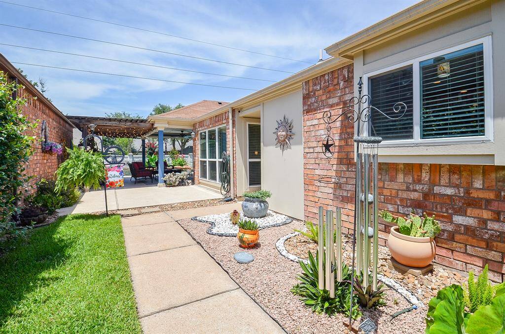 "Come on back and see," side yard, patio with a cozy seating area under a pergola, surrounded by vibrant plants and decorative accents. An inviting space for relaxation and outdoor entertaining.