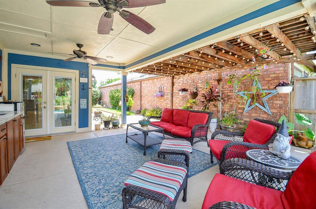 This inviting outdoor patio features ceiling fans and ambient string lights. The space is accented by a brick wall with decorative plants and a star, offering a cozy and stylish setting for relaxation or entertaining.