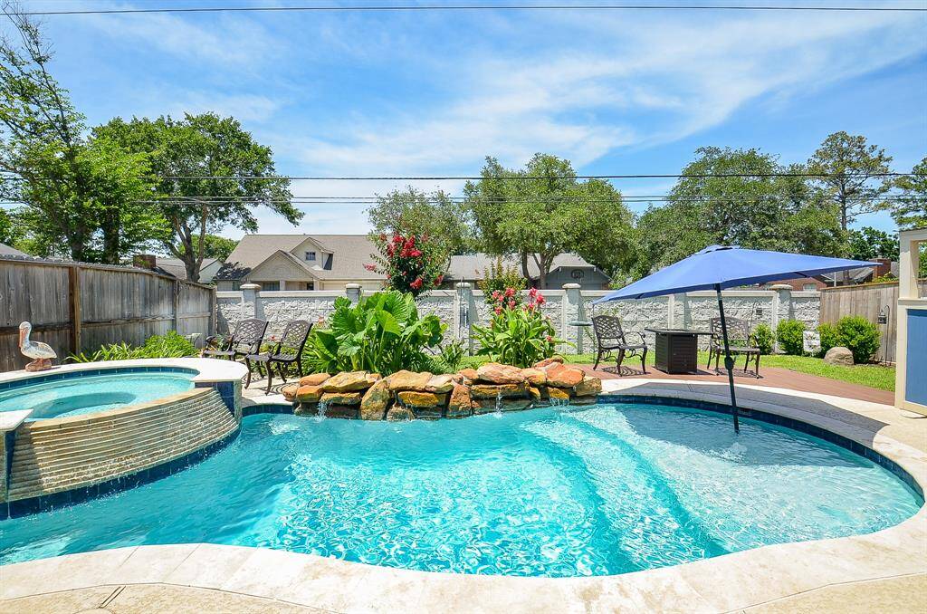 This backyard oasis features a sparkling pool with a stone waterfall, adjacent spa, and a cozy patio area with a large umbrella for shade. Surrounded by lush greenery and a privacy fence, it’s perfect for relaxation and entertaining.