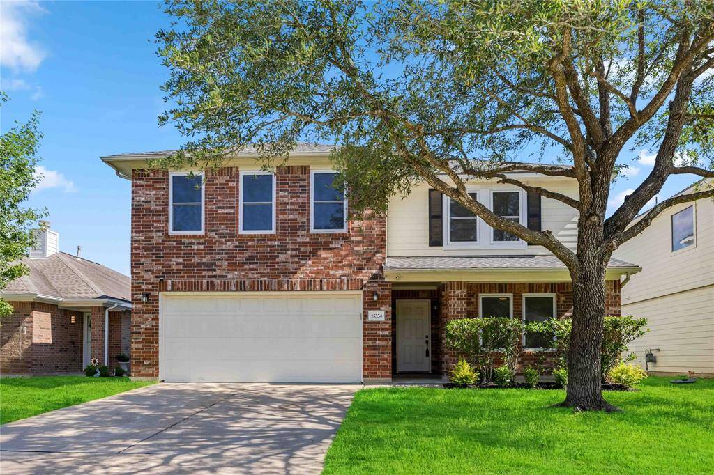 This inviting two-story home features a blend of brick and siding with a charming front porch, a two-car garage, and a well-maintained lawn accented by a mature tree.