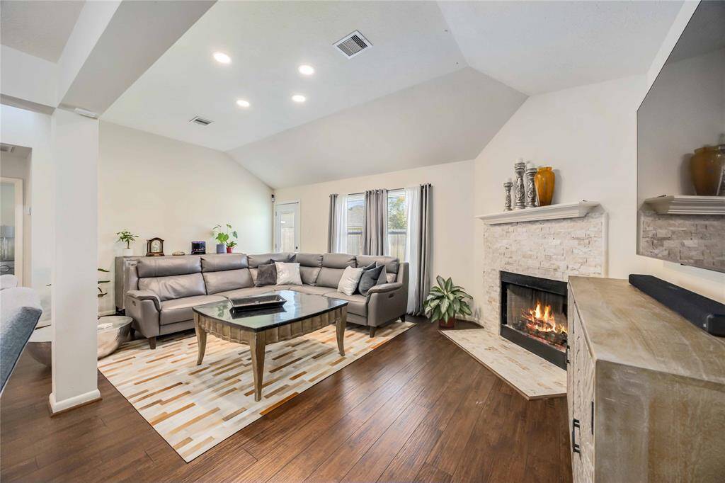 This inviting living room features a vaulted ceiling, a cozy fireplace, and hardwood flooring. A large sectional sofa and modern coffee table sit atop a stylish area rug, with ample natural light from the window. Perfect for relaxing and entertaining.