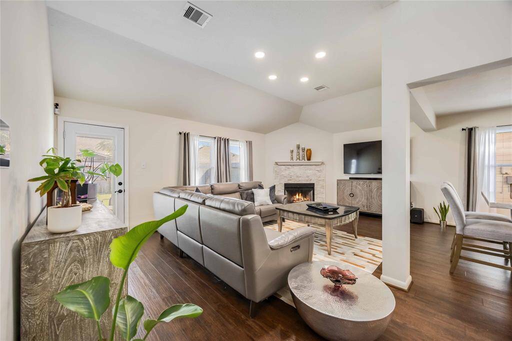 This inviting living room features a cozy fireplace, modern recessed lighting, and a spacious open layout. The room is accented with a stylish gray sectional sofa, wood flooring, and large windows that bring in natural light. A sleek entertainment setup and tasteful decor complete the contemporary feel.