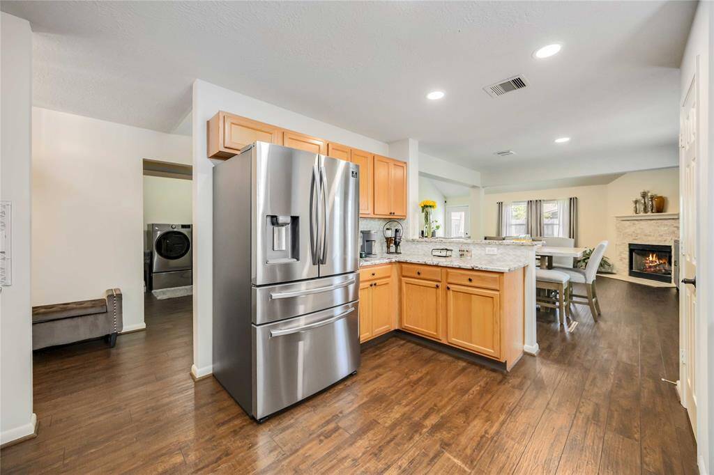 This inviting kitchen features stainless steel appliances, wooden cabinetry, and granite countertops. It seamlessly connects to a cozy dining area and living space with a fireplace, all set on beautiful hardwood floors.