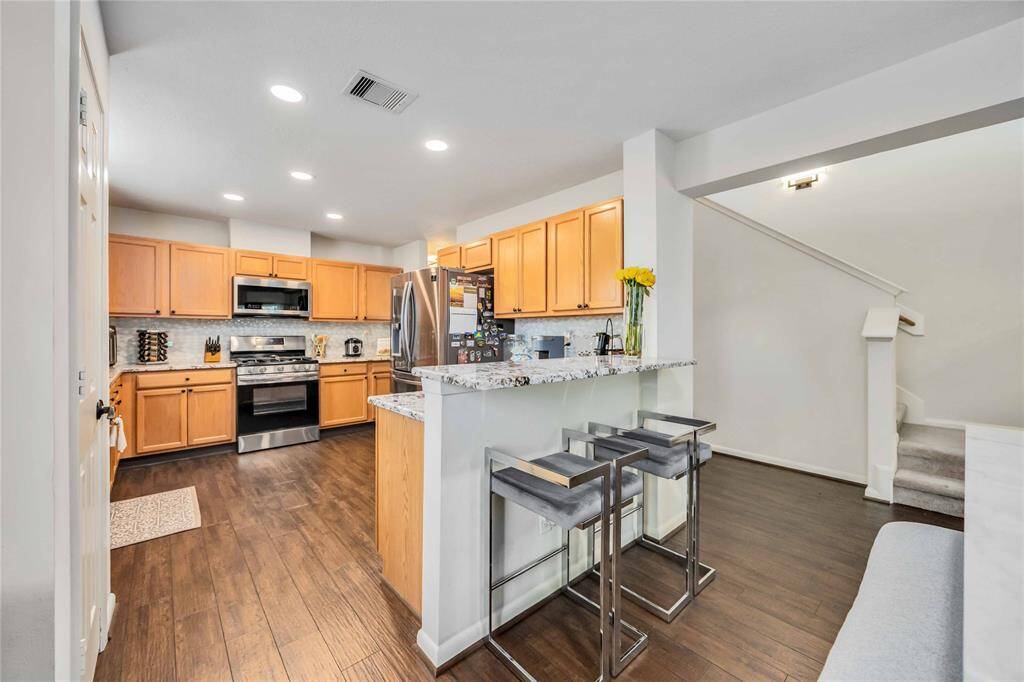 This photo showcases a modern kitchen with stainless steel appliances, light wood cabinetry, and a granite breakfast bar with seating. The space features recessed lighting and opens to a staircase, offering a clean and inviting layout ideal for entertaining.