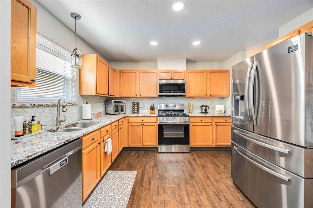 This modern kitchen features stainless steel appliances, warm wooden cabinets, and granite countertops. The space is well-lit with recessed lighting and a large window, complementing the stylish backsplash and hardwood floors.
