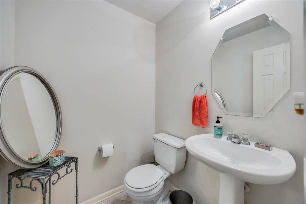 This is a compact, well-lit bathroom featuring a pedestal sink, a toilet, and an octagonal mirror. It includes a small decorative table with a round mirror and a bright accent towel.