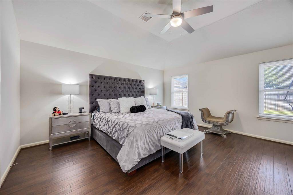 Spacious bedroom with a vaulted ceiling, featuring a plush, tufted headboard and stylish decor. It includes hardwood floors, ample natural light from large windows, and modern furniture, creating a comfortable and inviting atmosphere.