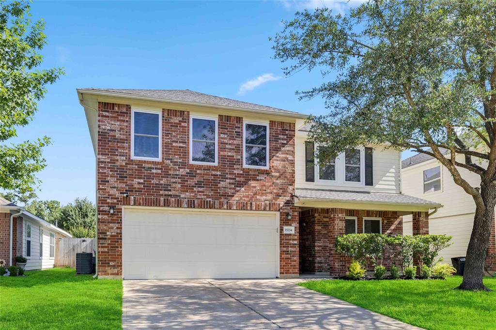 This is a two-story brick house with a two-car garage and a neatly maintained front yard. It features a covered entryway with shrubs and a large tree providing shade. The exterior is a mix of brick and siding, giving it a classic and inviting look.