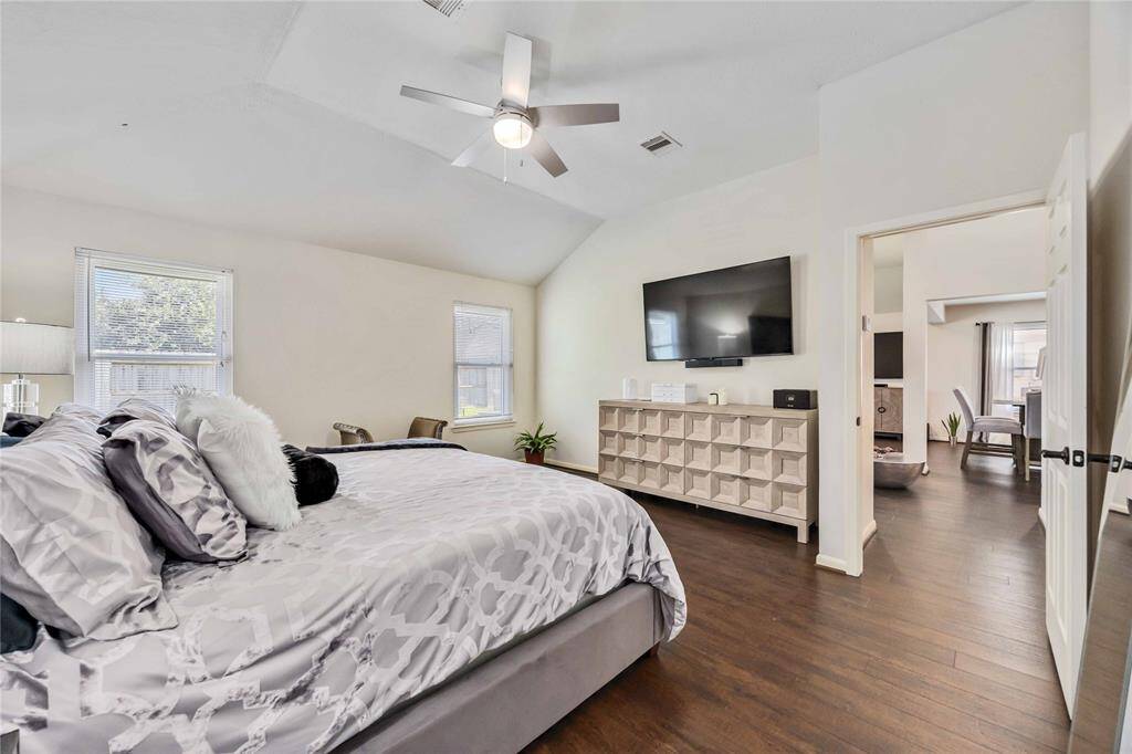 Spacious bedroom with vaulted ceilings, hardwood floors, and ample natural light. Features a ceiling fan, mounted TV, and stylish furniture. Opens to a living area, creating a seamless flow. Ideal for comfort and relaxation.