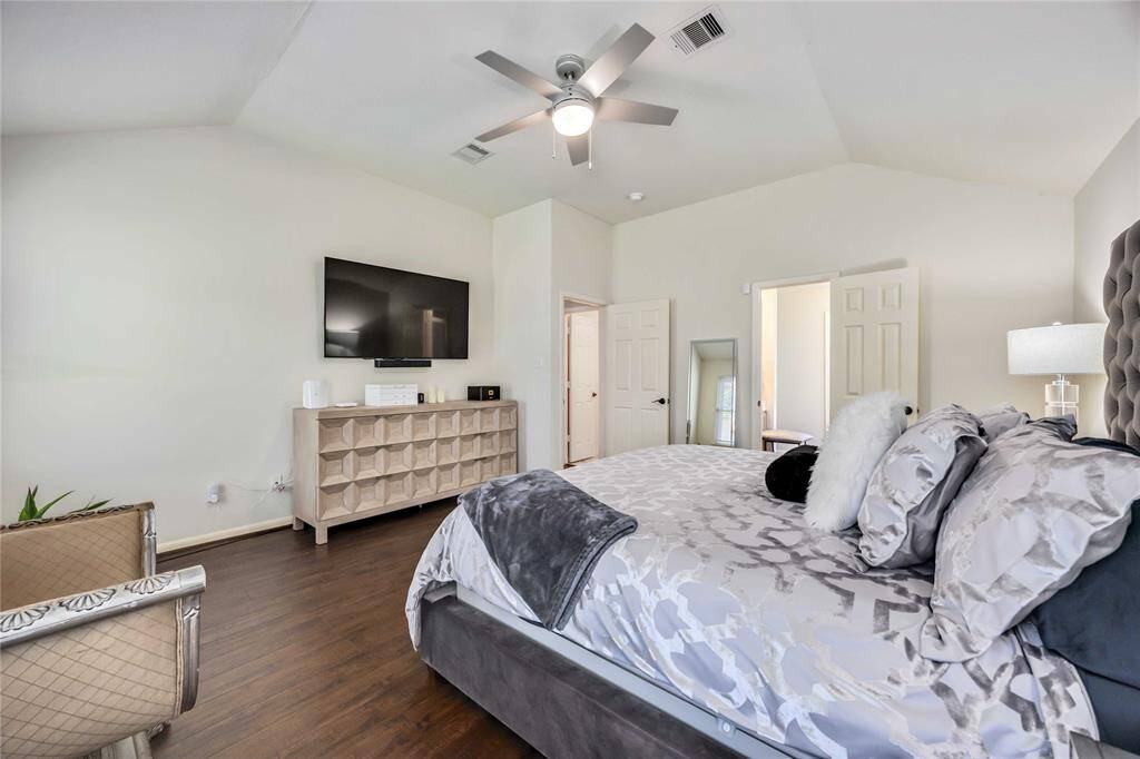 This spacious bedroom features a sleek modern design with hardwood floors, a ceiling fan, and a wall-mounted TV. It includes a comfortable bed with plush bedding, ample natural light, and a stylish dresser, creating a cozy and inviting atmosphere.
