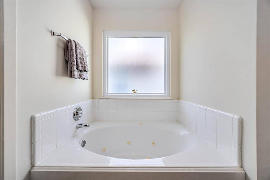 This photo features a clean, white-tiled bathroom with a spacious jetted bathtub and a frosted window above, allowing natural light while maintaining privacy. A towel rack is conveniently located nearby.