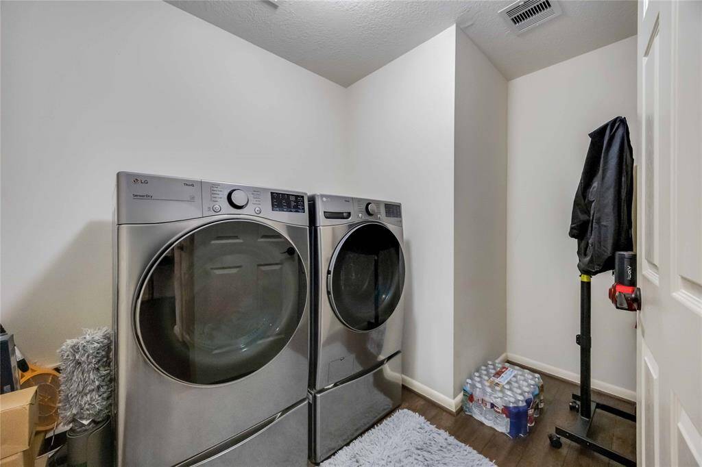 This photo shows a laundry room with modern front-loading washer and dryer units. The room has white walls, wooden flooring, and a small rug. Storage items are tucked neatly, and there's additional space for supplies.