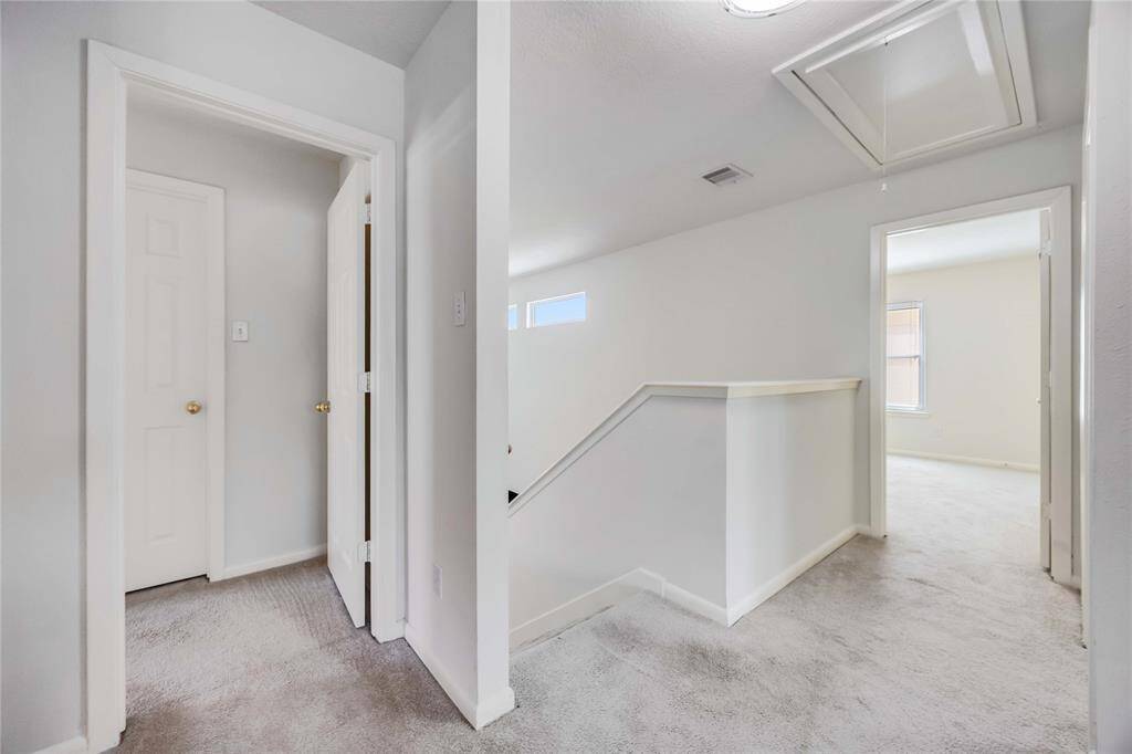This photo shows a bright, carpeted upstairs hallway with white walls. It features two doorways leading to separate rooms and a stairway with a half-wall railing. The area is well-lit with natural light.