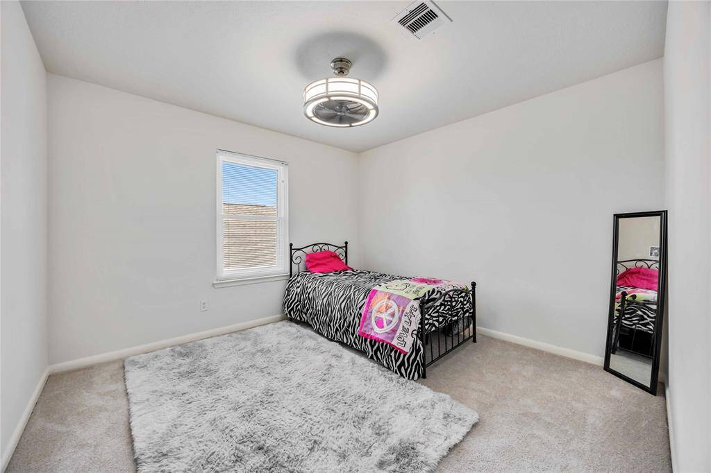 This cozy bedroom features neutral walls, a modern ceiling light, and a single window for natural light. It includes a twin bed with a colorful bedspread, a plush area rug, and a full-length mirror, creating a welcoming space.