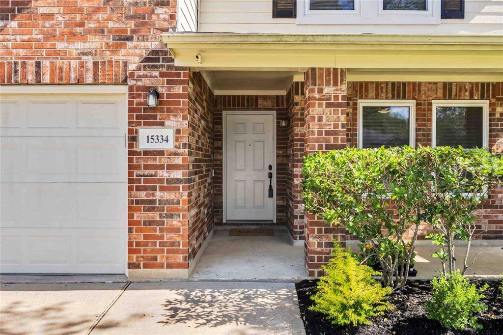 Charming brick exterior with a covered entryway, featuring a white front door and adjacent garage. Well-maintained shrubs add curb appeal.