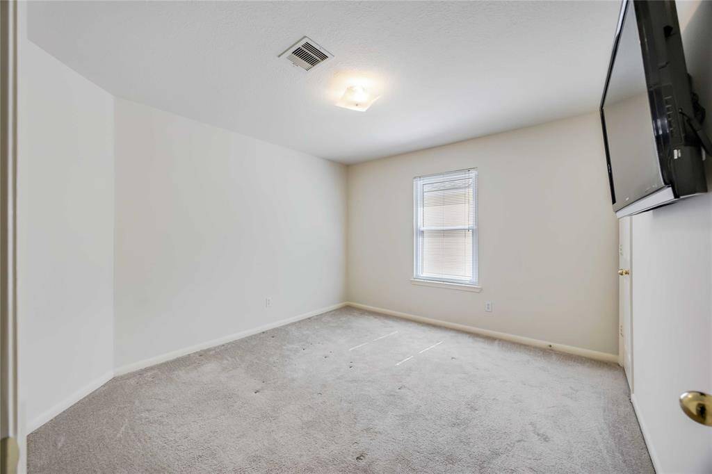 This photo showcases a simple, unfurnished bedroom with neutral walls, carpeted flooring, a ceiling light, and a wall-mounted television. A window allows natural light to brighten the space.