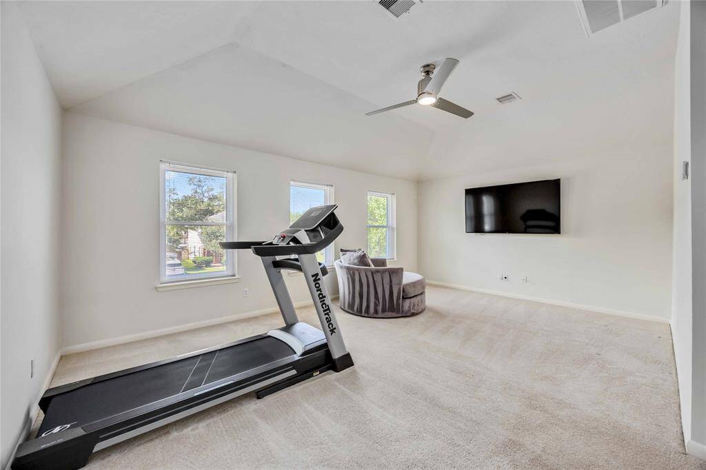 This spacious, well-lit room features plush carpeting and large windows. It includes a mounted TV, a treadmill, and a cozy seating area with a ceiling fan for comfort. Ideal for a home gym or relaxation space.