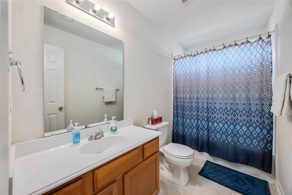 This bathroom features a clean and modern design with a large mirror, ample counter space, and a wooden vanity. It includes a toilet and a shower with a stylish blue and white patterned curtain. Bright lighting and neutral tones create a fresh and inviting atmosphere.