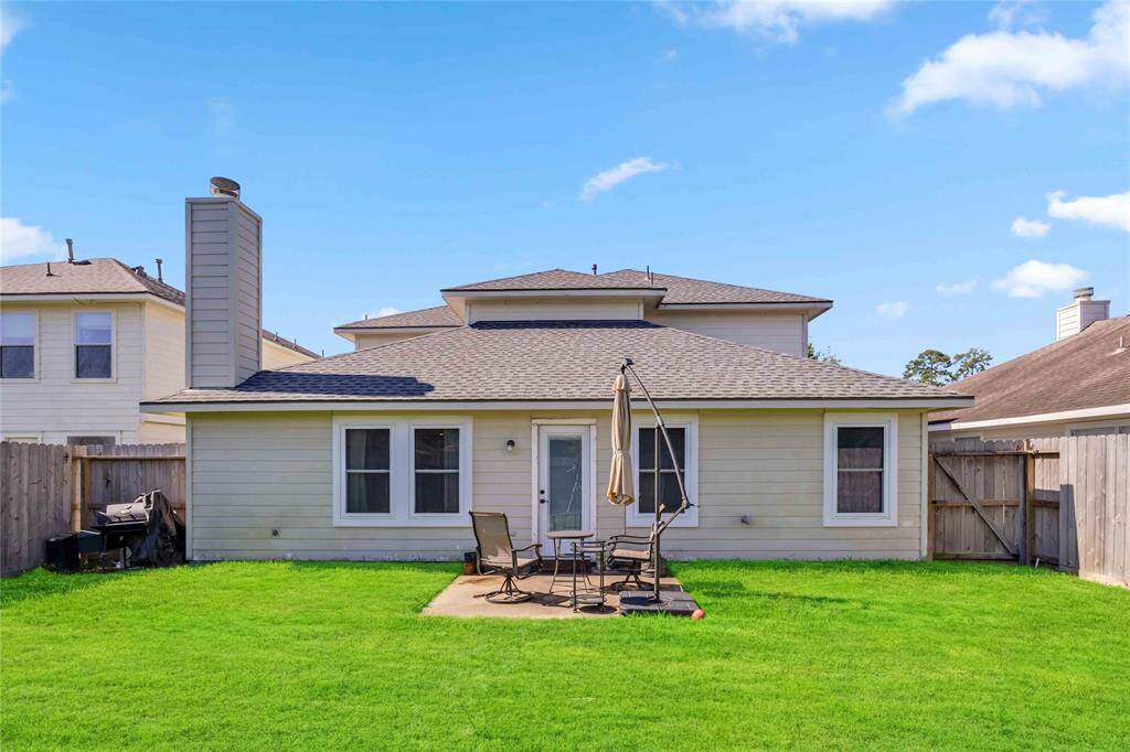 This photo shows a backyard with a well-maintained lawn, a small patio area featuring outdoor furniture and an umbrella, a barbecue grill, and a wooden fence for privacy. The home's exterior is light-colored with a shingled roof.