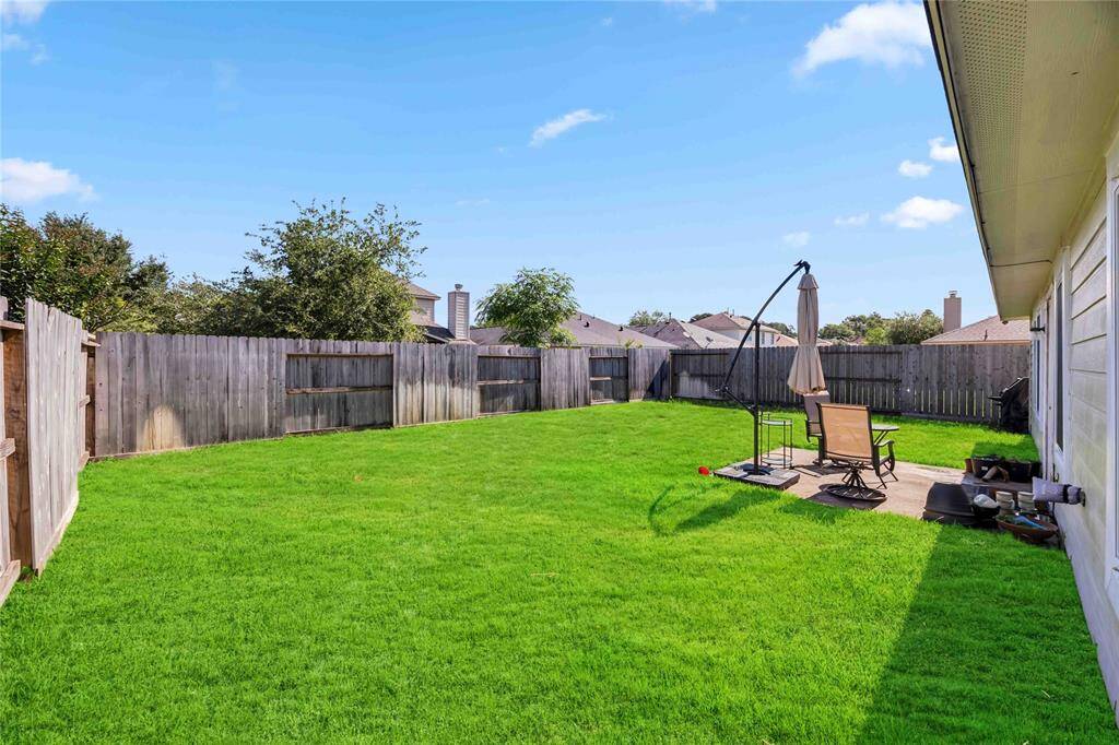 Spacious backyard with lush green lawn, enclosed by a wooden fence. Features a small patio area with seating and an umbrella, ideal for outdoor relaxation.