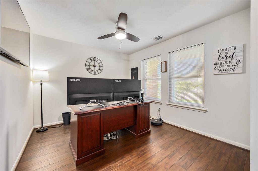This home office features a spacious layout with hardwood floors, ample natural light from two large windows, a ceiling fan, and a modern desk setup with dual monitors. Ideal for productivity and comfort.