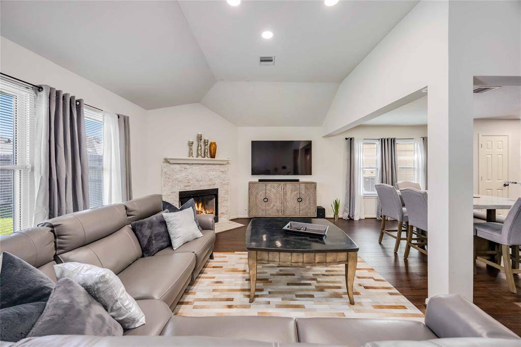 Spacious living room with high ceilings and abundant natural light. Features include a cozy fireplace, elegant wood flooring, and a modern open layout. The room is tastefully furnished with a large sectional sofa, stylish coffee table, and a wall-mounted TV, leading into a dining area.