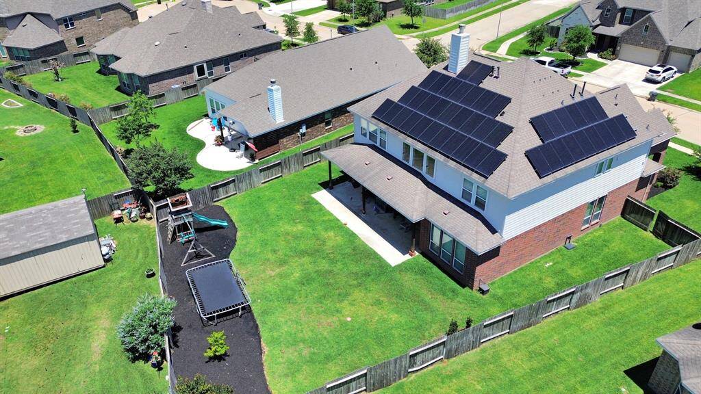 This aerial view showcases a spacious two-story home with solar panels on the roof, a large fenced backyard, and a well-maintained lawn. The yard features a trampoline, playground set, and a patio area, offering ample outdoor space for family activities.