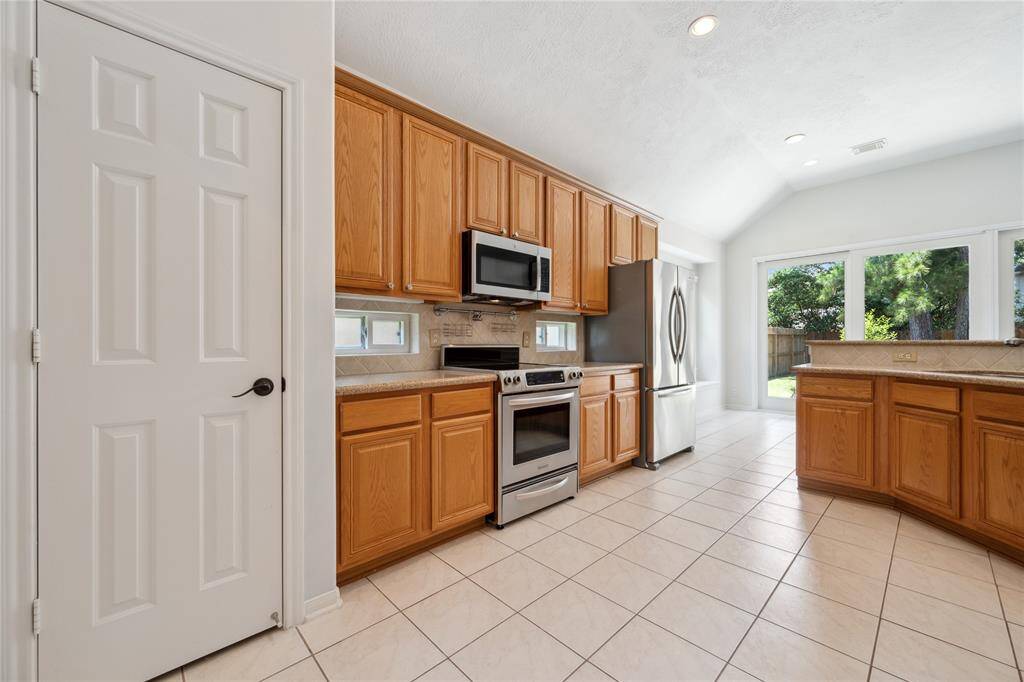 This kitchen features ample natural light, modern stainless steel appliances, and warm wooden cabinetry. The open layout includes tile flooring and a view of the backyard through large windows, creating an inviting and functional space for cooking and dining.