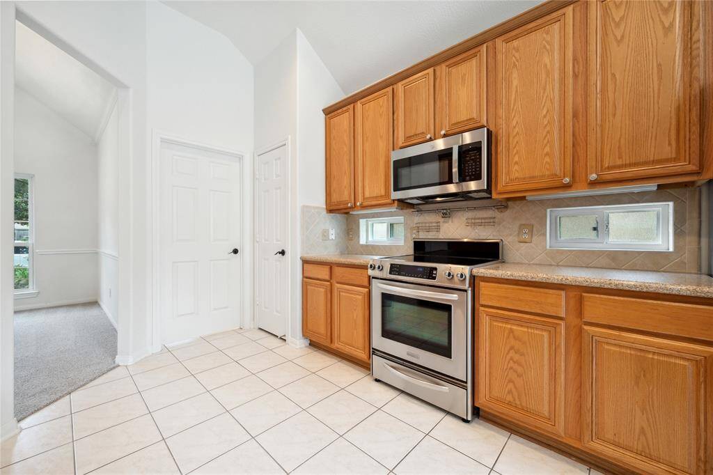 This kitchen features stainless steel appliances, wooden cabinetry, and tile flooring. It has a modern microwave and oven, ample storage, and small windows for natural light. The adjacent hallway leads to a carpeted room.