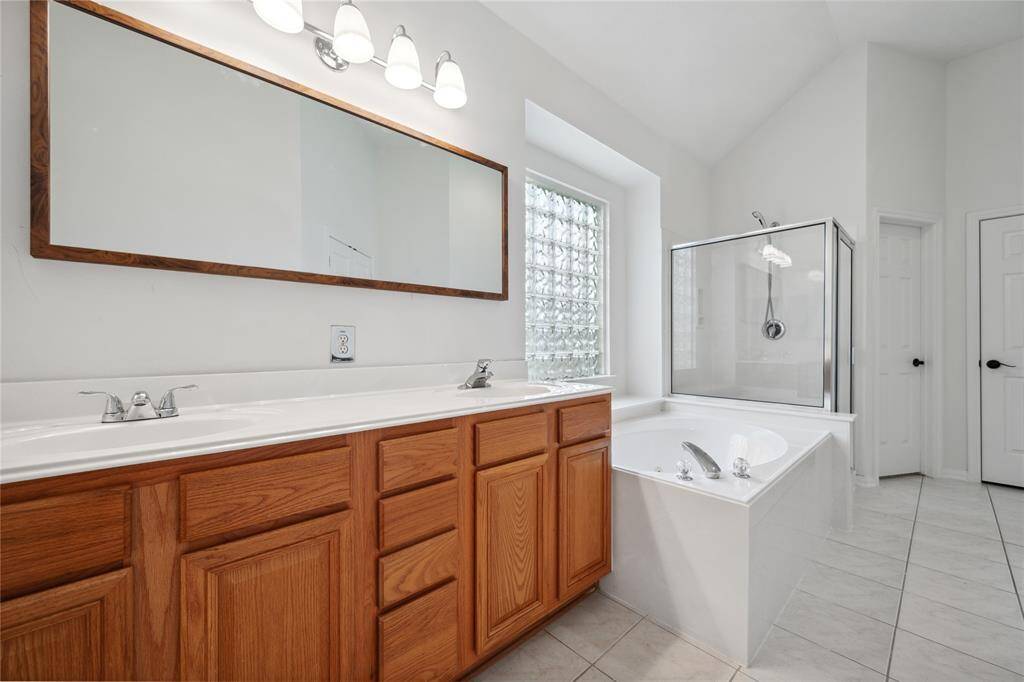 This bathroom features a spacious double vanity with wooden cabinets, a large mirror, and modern lighting. It has a separate glass-enclosed shower and a soaking tub, complemented by neutral tiles and natural light from a glass block window.