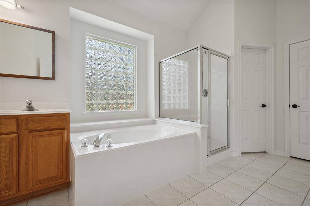 Bright and spacious bathroom featuring a large soaking tub, separate glass-enclosed shower, and a wooden vanity with a mirror. The space is enhanced by a glass block window, providing natural light and privacy.