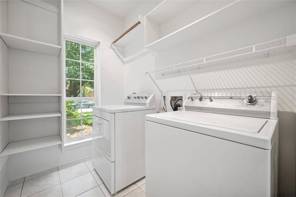 Bright laundry room with a large window, featuring a washer and dryer, ample shelving, and tiled flooring.