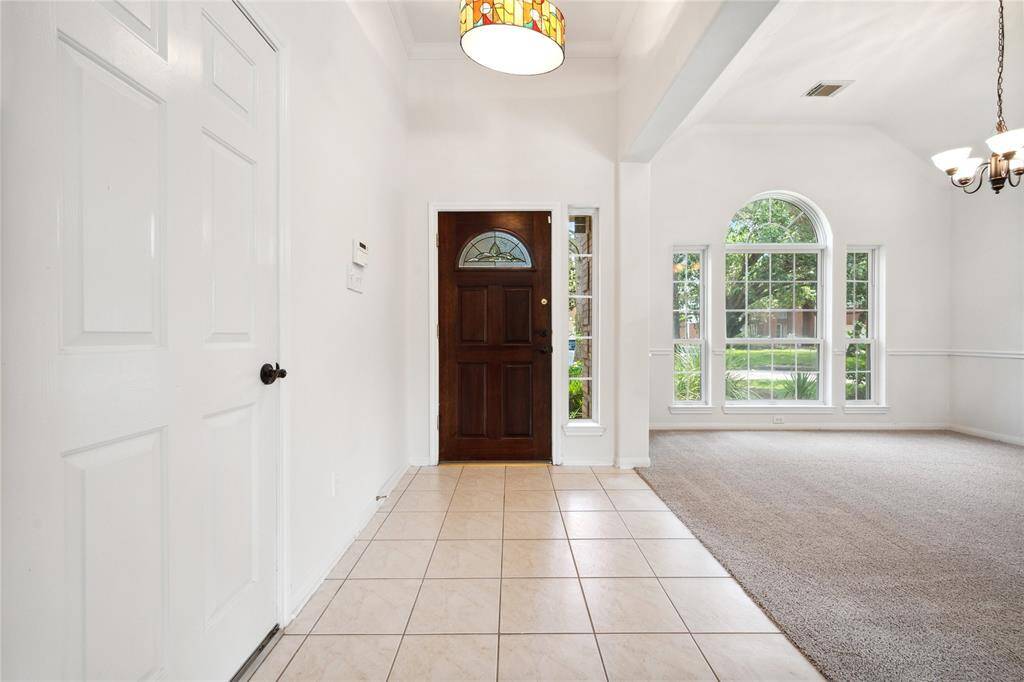 Bright entryway with a warm wooden door, tiled flooring, and a spacious adjoining carpeted room featuring large arched windows and a chandelier.